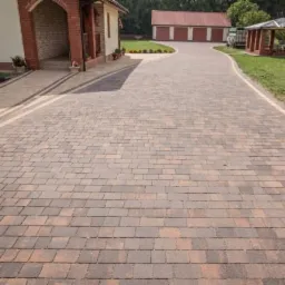 Brukowana droga dojazdowa z kostki w odcieniach brązu prowadząca do domu z ceglanym łukiem wejściowym, w tle garaż z czerwonym dachem i altana.