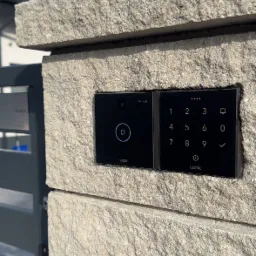 Zewnętrzny panel dotykowy Loxone z kamerą, zamontowany na kamiennym słupku ogrodzenia posesji.