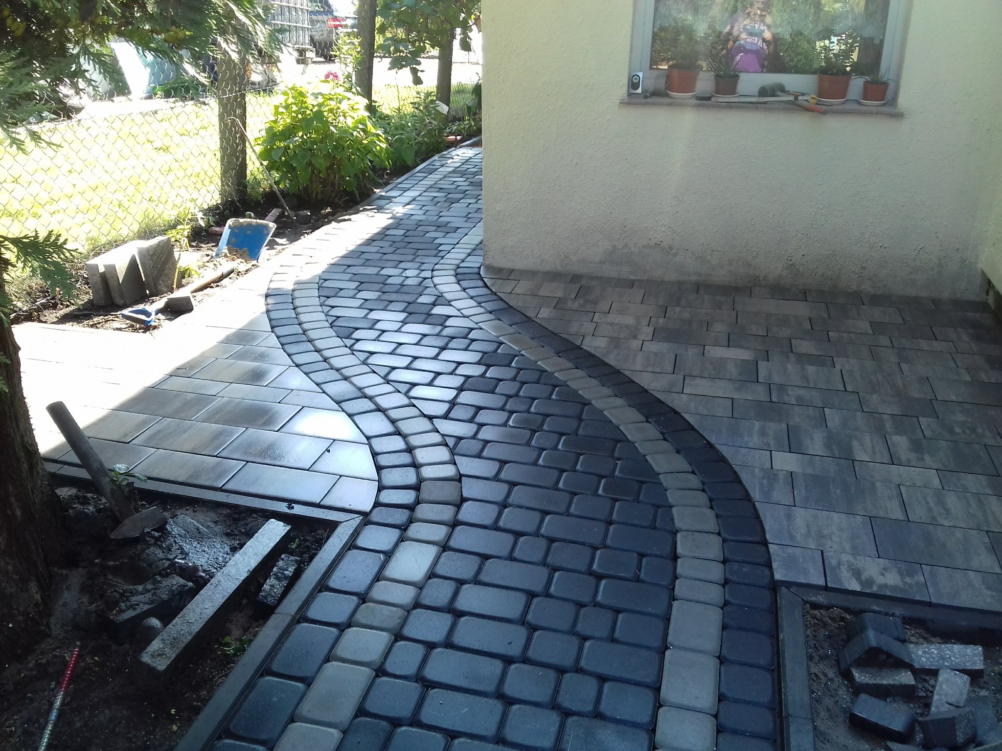 Ścieżka z kostki granitowej w dwóch odcieniach szarości, ułożona w łukowaty wzór obok budynku z kwiatami w oknie i narzędziami w pobliżu drzewa.