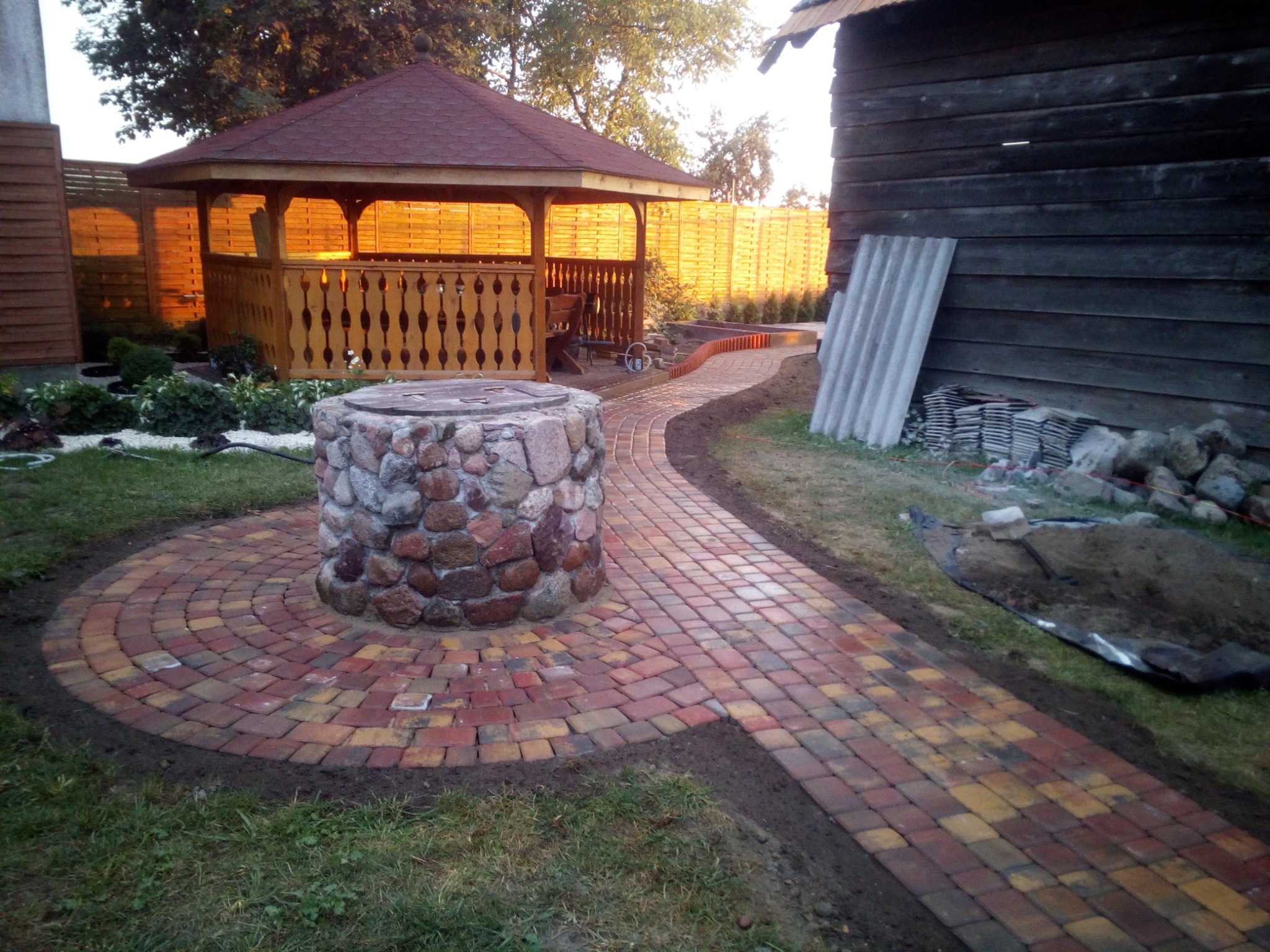 Ogród z altaną i ścieżką z kostki, studnia obłożona kamieniami. Ścieżka prowadzi do altany, w tle drewniany płot i budynek. Widoczne elementy dekoracyjne i roślinność.