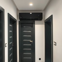DMS System sp.z o.o. - Nowoczesny czarny klimatyzator zamontowany nad drzwiami w korytarzu, zabezpieczony kartonami na podłodze podczas remontu.