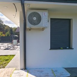 DMS System sp.z o.o. - Biała jednostka zewnętrzna klimatyzacji Rotenso Teta Series zamontowana na ścianie budynku z roletą zewnętrzną, zabezpieczona kartonami na trawniku.