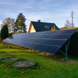 DMS System sp.z o.o. - Panel fotowoltaiczny zamontowany na metalowej konstrukcji w ogrodzie, w tle dom z panelami słonecznymi na dachu i jesienne drzewa pod błękitnym niebem.