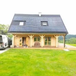 Drewniany dom z panelami fotowoltaicznymi na dachu, widoczny samochód dostawczy i narzędzia sugerujące trwającą instalację.