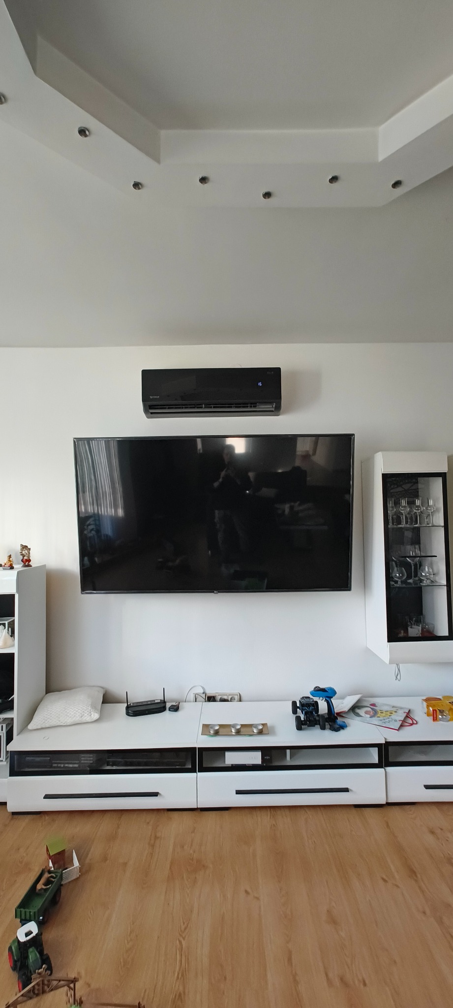 Czarny klimatyzator ścienny zamontowany nad telewizorem w salonie, widoczny fragment sufitu z oświetleniem punktowym i białe meble.