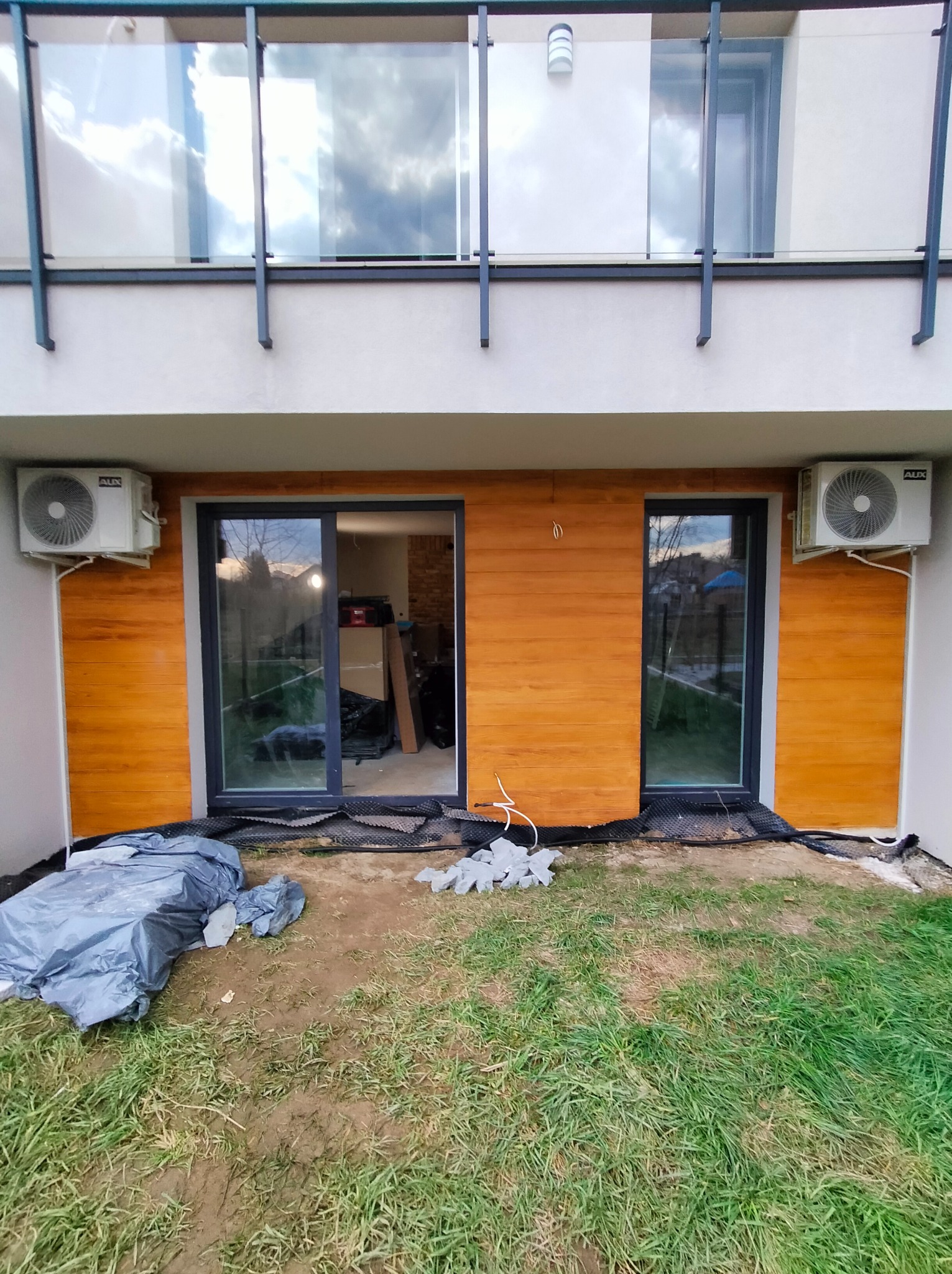 Dwa klimatyzatory zamontowane na elewacji budynku jednorodzinnego, po obu stronach przeszklonych drzwi tarasowych. Na trawniku widoczne ślady prac instalacyjnych, zabezpieczone folią i matą.