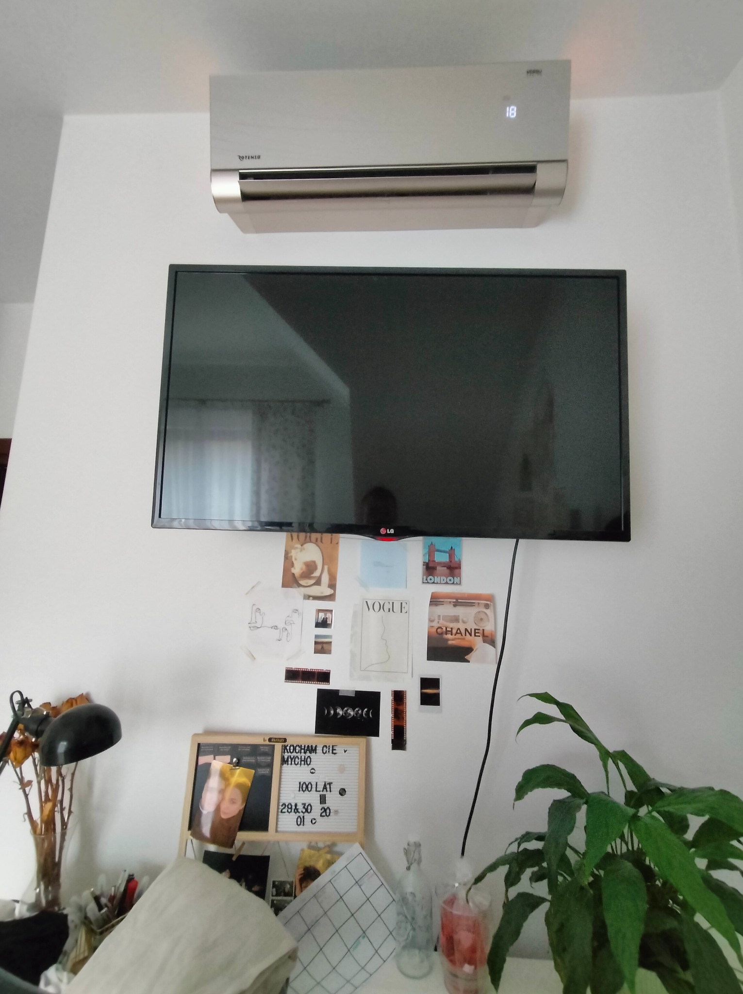 Nowoczesny klimatyzator na ścianie nad telewizorem, pod nim kolaż zdjęć i dekoracji na białej ścianie w pokoju.