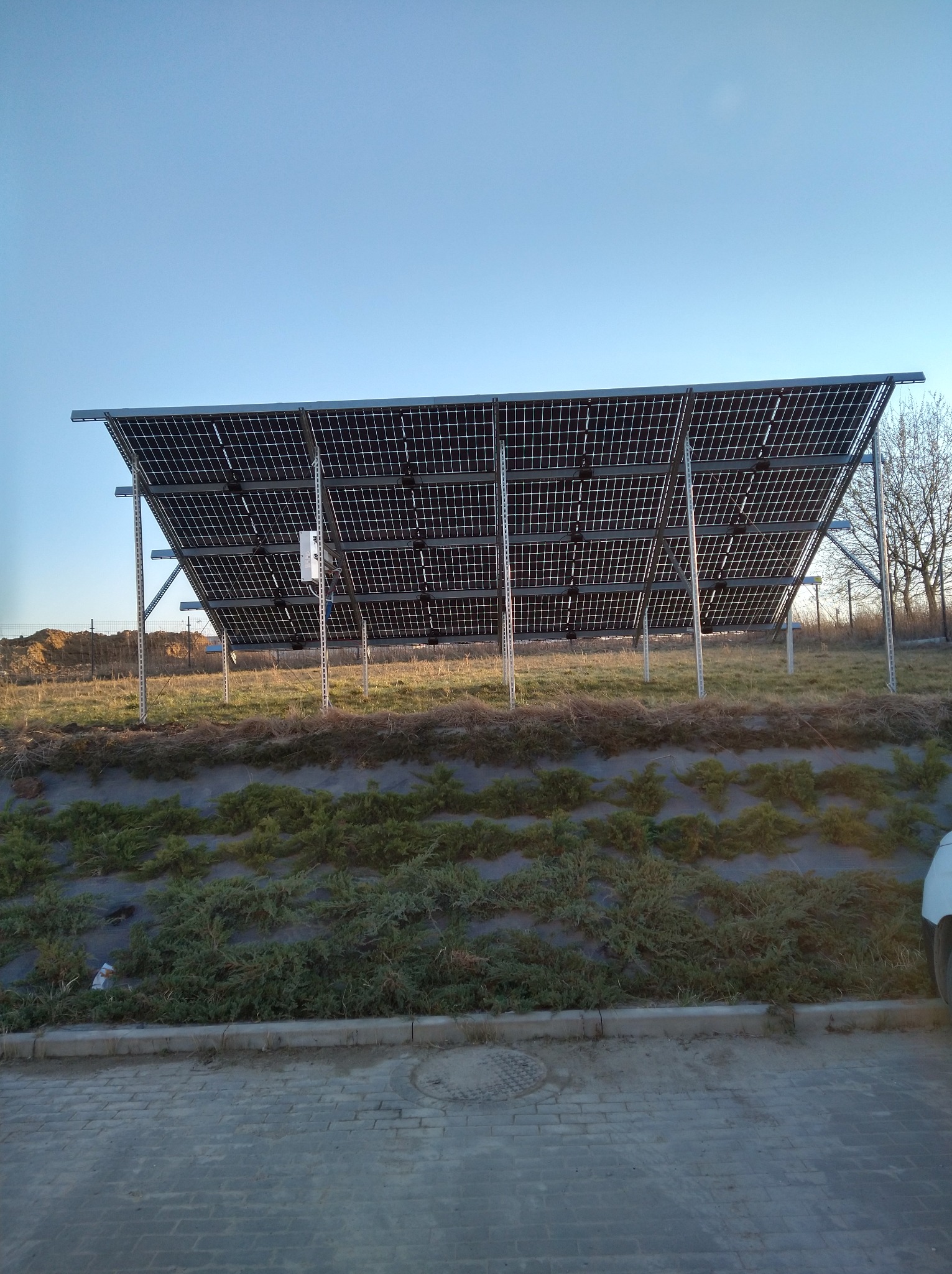 Instalacja paneli fotowoltaicznych na metalowej konstrukcji, widok od tyłu, z niską roślinnością w tle i fragmentem chodnika z kostki brukowej w dolnej części kadru.