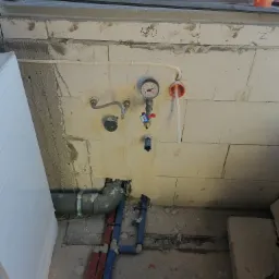 Surowy stan instalacji wodno-kanalizacyjnej w trakcie montażu: widoczne rury, manometr, przyłącza, fragment okna i ściany z betonu kom&oacute;rkowego z oznaczeniami.