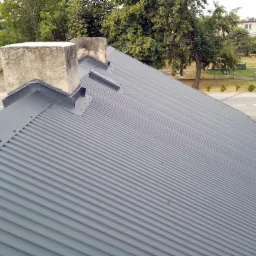 Szary dach z blachy trapezowej po renowacji, widoczne dwa kominy z betonowymi czapami i elementy obróbki blacharskiej, w tle drzewa i zabudowania.