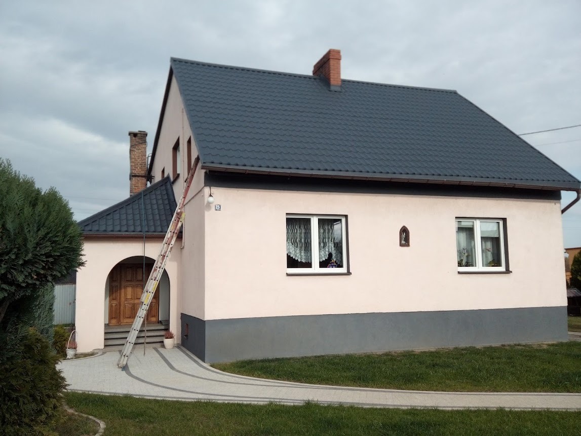 Dwupiętrowy dom z szarym dachem z dachówki, jasnoróżową elewacją i drewnianymi drzwiami wejściowymi z łukowym przejściem. Do ściany oparta drabina. Widoczne okna z firankami i mała kapliczka...