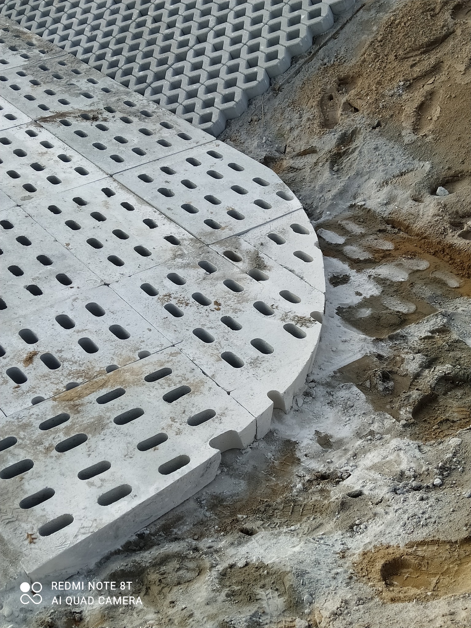 Fragment powierzchni z ażurowych płyt betonowych z owalnymi otworami, układanych na piasku, widoczne ślady pracy i przygotowania podłoża.