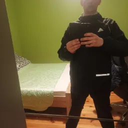 Mężczyzna w czarnej kurtce Adidas robi sobie selfie w lustrze; widoczny fragment pokoju z zieloną ścianą i łóżkiem.