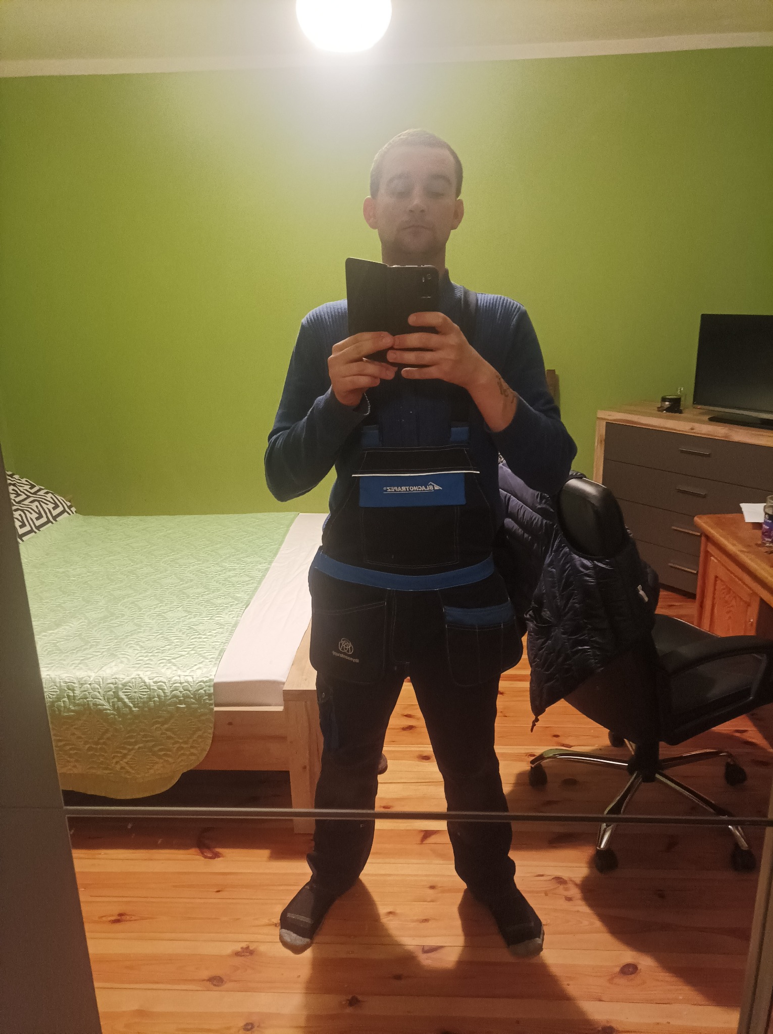 Mężczyzna w roboczym kombinezonie, robiący sobie selfie w lustrze w pokoju z zieloną ścianą i drewnianą podłogą.