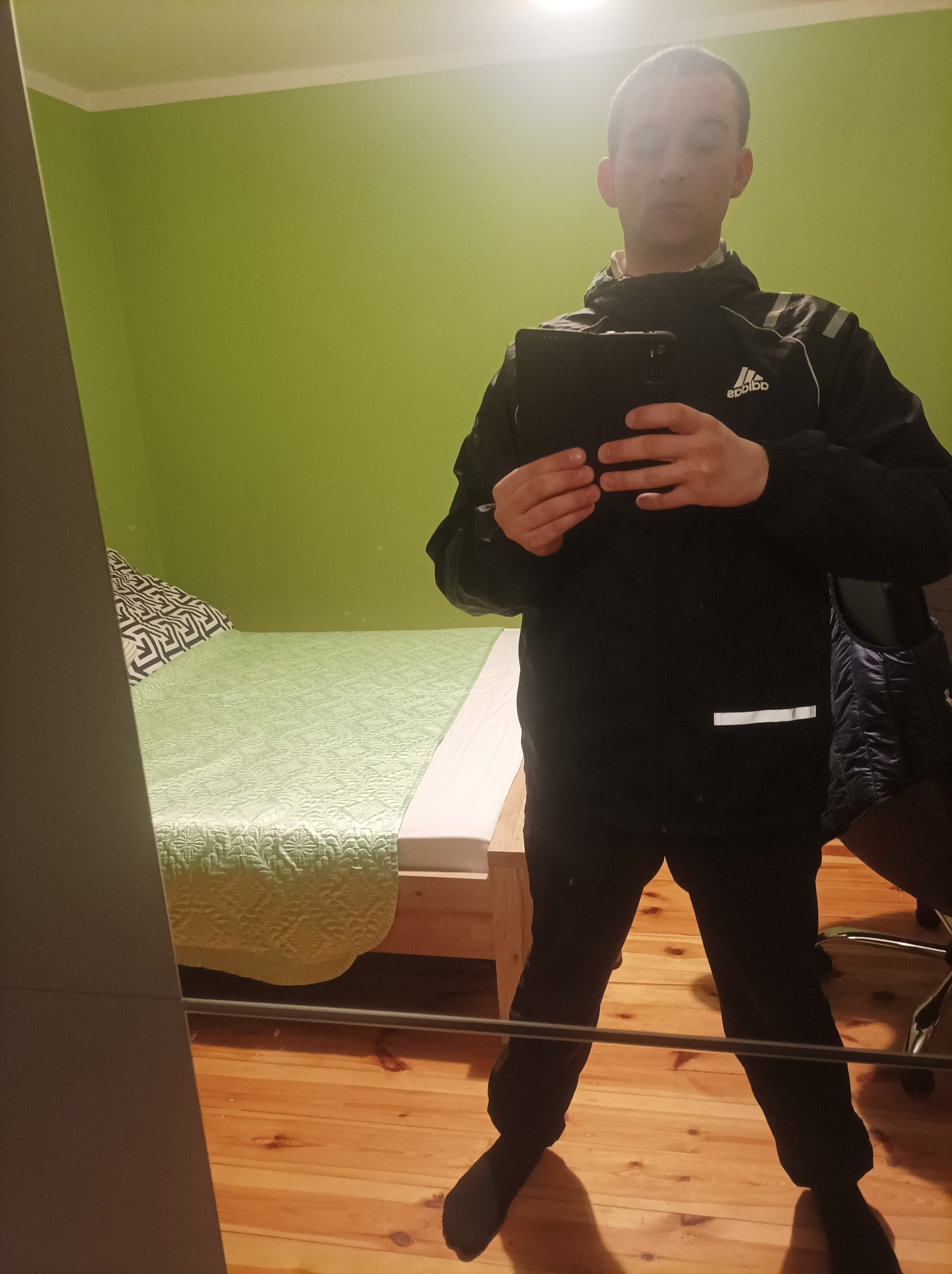Mężczyzna w czarnej kurtce Adidas robi sobie selfie w lustrze; widoczny fragment pokoju z zieloną ścianą i łóżkiem.