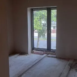 Wnętrze pokoju w trakcie prac wykończeniowych: ściany pokryte świeżym tynkiem, widoczne ślady po pracach na podłodze zabezpieczonej listwami, okno z widokiem na ulicę i drzewa.