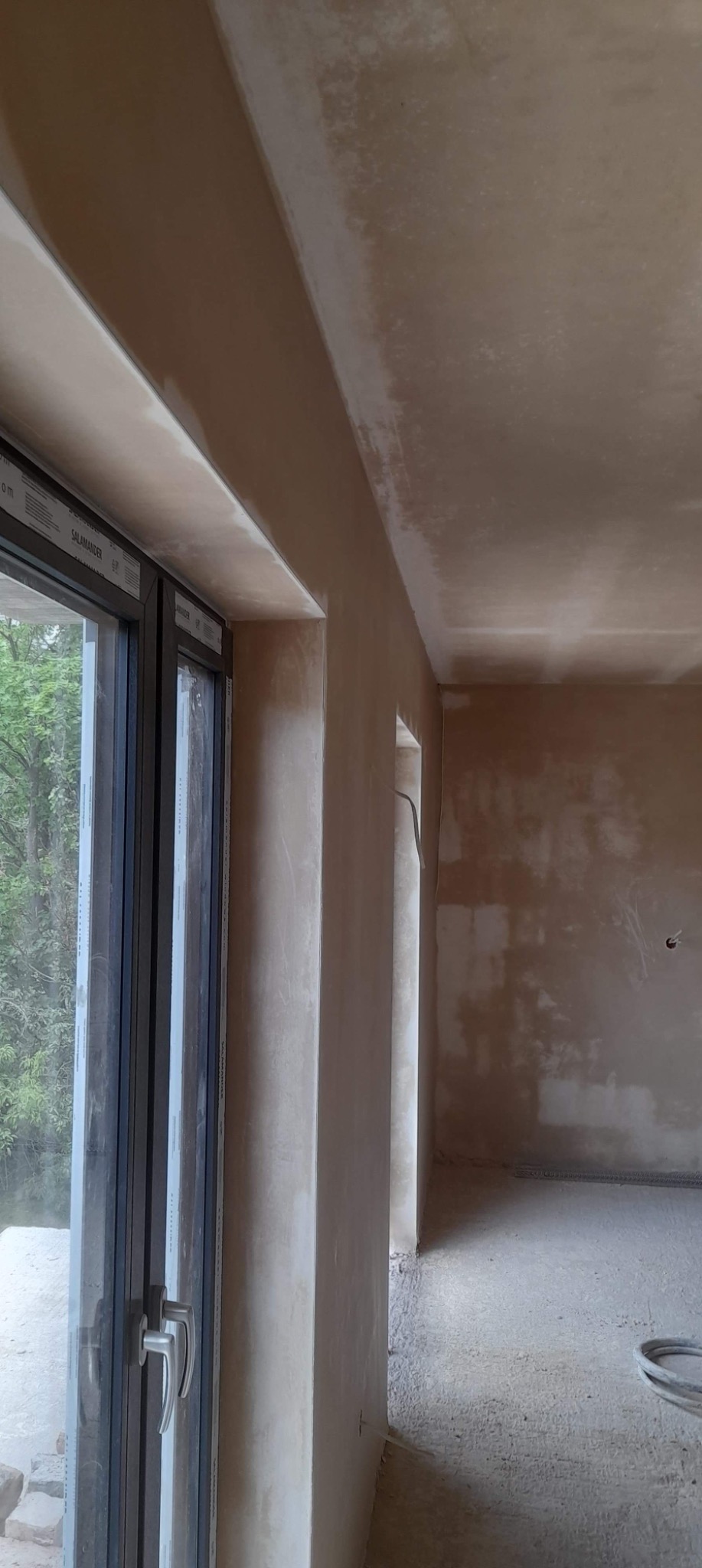 Wnętrze w trakcie prac wykończeniowych, widoczna ściana po nałożeniu tynku, okno z widokiem na zieleń na zewnątrz, surowa podłoga, ślady po instalacjach na ścianie.