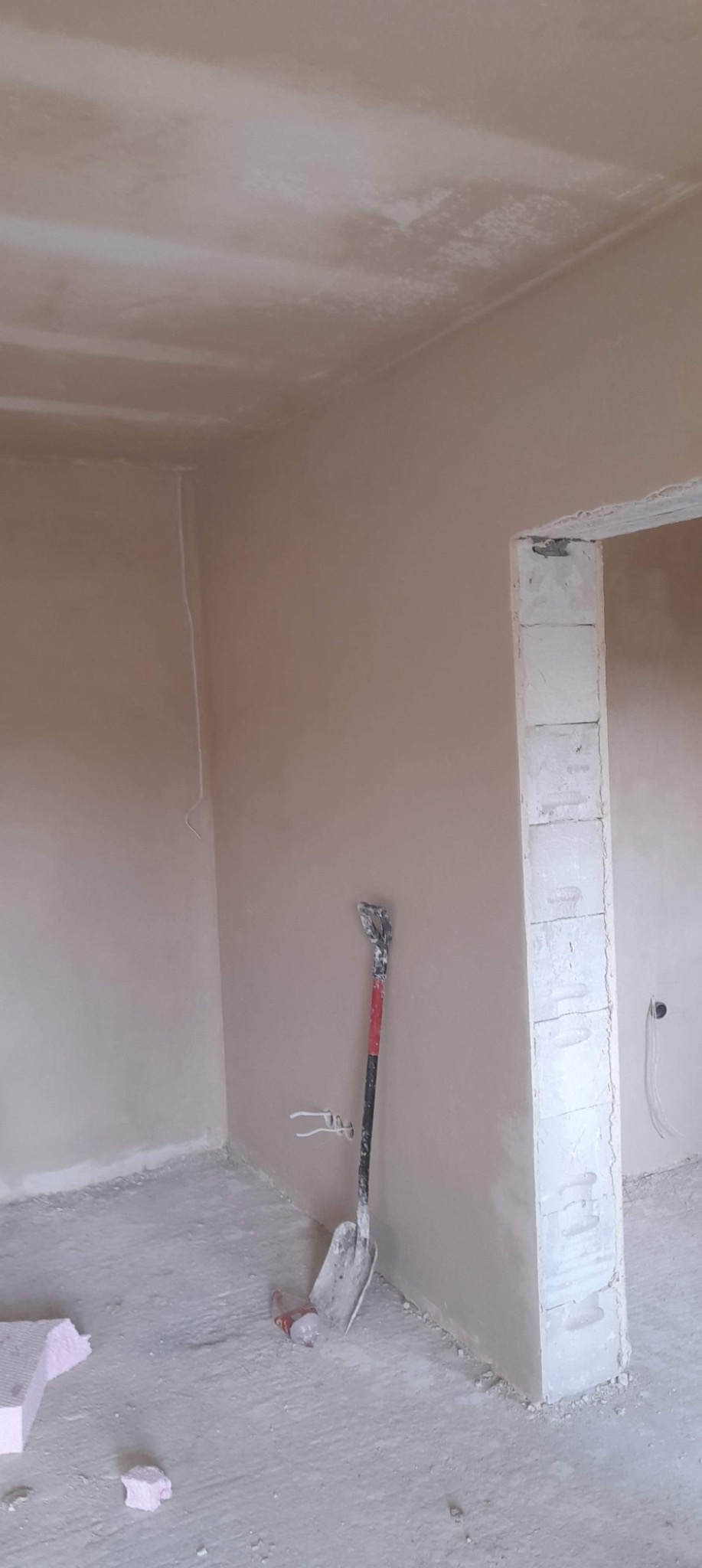 Wnętrze w trakcie prac wykończeniowych: ściany pokryte świeżym tynkiem, widoczna łopata oparta o ścianę, fragmenty styropianu na podłodze, surowy otwór drzwiowy z bloczków.