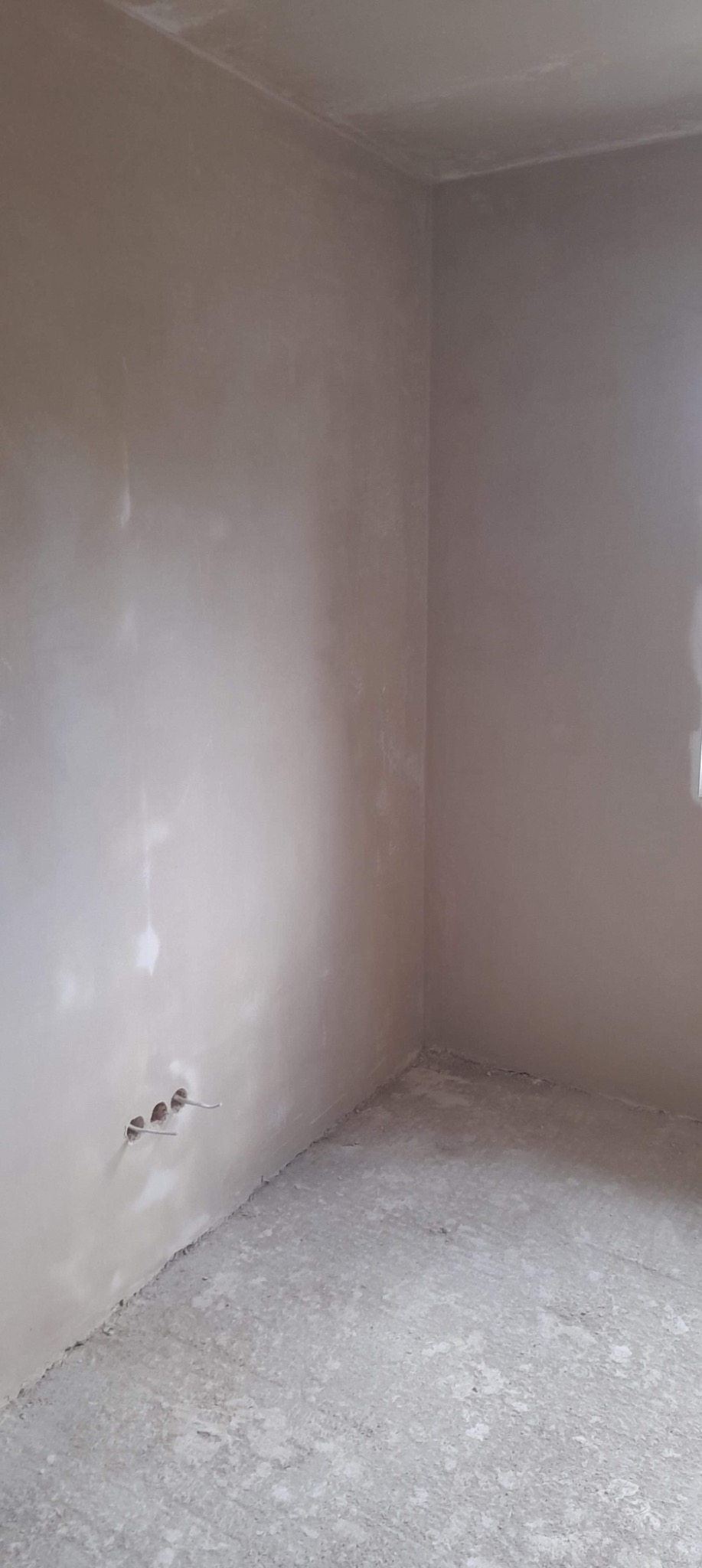 Wnętrze pokoju po nałożeniu jasnoszarej gładzi szpachlowej na ściany i sufit, widoczne wystające kable elektryczne ze ściany na dole po lewej stronie, surowy beton na podłodze.