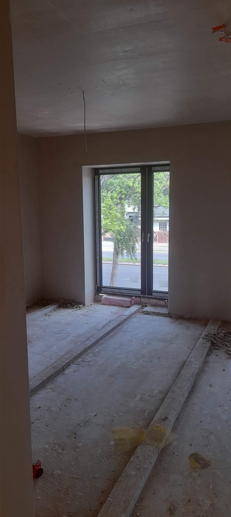Wnętrze pokoju w trakcie prac wykończeniowych: ściany pokryte świeżym tynkiem, widoczne ślady po pracach na podłodze zabezpieczonej listwami, okno z widokiem na ulicę i drzewa.