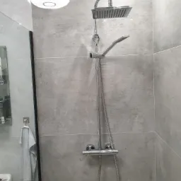 Nowoczesny zestaw prysznicowy z deszczownicą i słuchawką, zamontowany na szarej, imitującej kamień ścianie w łazience. Widoczny termostat i wąż prysznicowy. W tle fragment lustra i biały ręcznik.