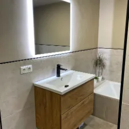 Nowoczesna łazienka z podświetlanym lustrem LED, umywalką nablatową z szafką z drewnopodobnym dekorem, czarną armaturą i płytkami w odcieniach szarości.