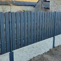 Nowoczesne, szare ogrodzenie panelowe z podmurówką imitującą kamień, widoczne na tle budynku z cegły i drzew bez liści.