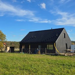 CONPEKA Piotr Krukierek - Nowoczesny, czarny dom szkieletowy z ciemnym dachem na tle zielonej łąki i błękitnego nieba. Widoczne samochody dostawcze i drzewo z żółtymi liśćmi.