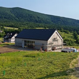 CONPEKA Piotr Krukierek - Wznoszenie domu szkieletowego na tle zalesionych wzgórz. Budynek w stanie surowym zamkniętym, z widocznymi rusztowaniami i elementami konstrukcyjnymi.