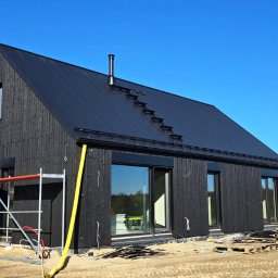 CONPEKA Piotr Krukierek - Nowoczesny dom szkieletowy z czarną elewacją i dachem na tle błękitnego nieba. Widoczne rusztowania i elementy wykończeniowe.