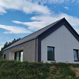 CONPEKA Piotr Krukierek - Nowoczesny dom jednorodzinny z dwuspadowym dachem, wykończony w odcieniach szarości i bieli, usytuowany na wzgórzu z widokiem na zalesione tereny w tle.