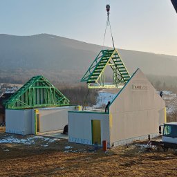 CONPEKA Piotr Krukierek - Montaż domu modułowego z użyciem dźwigu na tle górskim. Widoczne elementy konstrukcyjne i logo producenta na ścianie. Jasne, poranne światło.