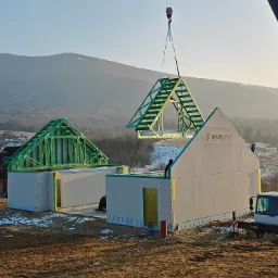 Montaż domu modułowego z użyciem dźwigu na tle górskim. Widoczne elementy konstrukcyjne i logo producenta na ścianie. Jasne, poranne światło.