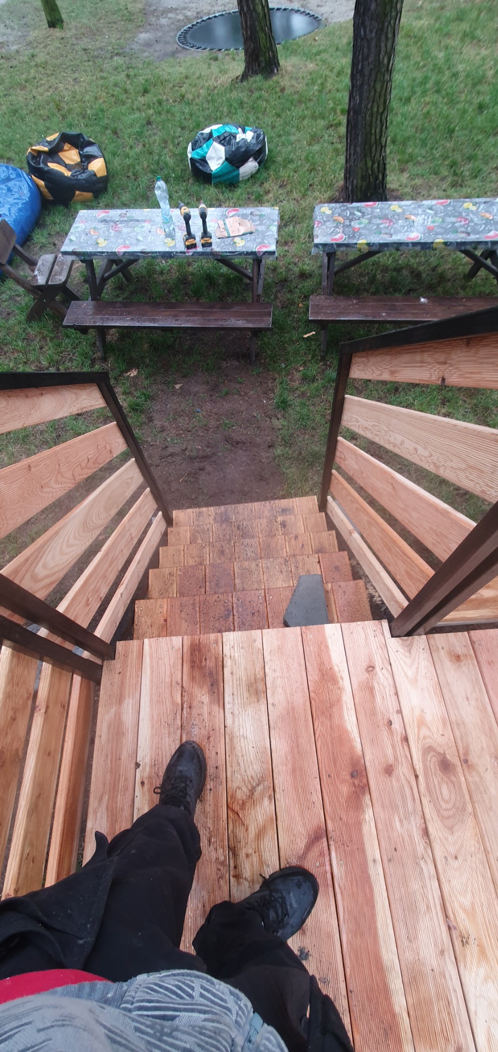 Widok z góry na drewniane schody zewnętrzne z barierkami, prowadzące w dół, z widocznymi butami osoby stojącej na górnym stopniu; w tle stoliki piknikowe, pufy i trampolina.