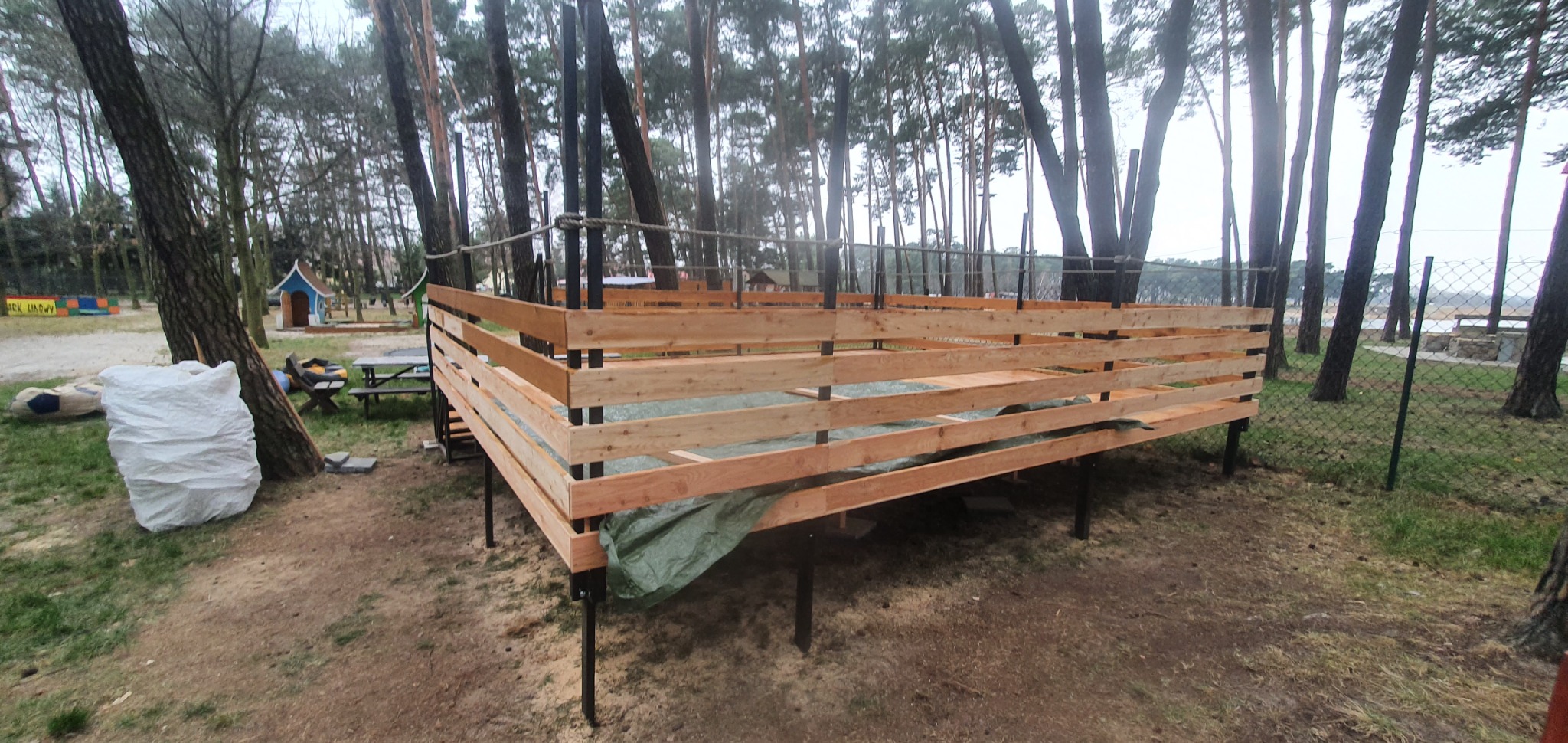 Konstrukcja z surowego drewna na metalowych podporach, prawdopodobnie taras, w otoczeniu drzew iglastych i z widokiem na wodę w tle. Przykryta zieloną plandeką.