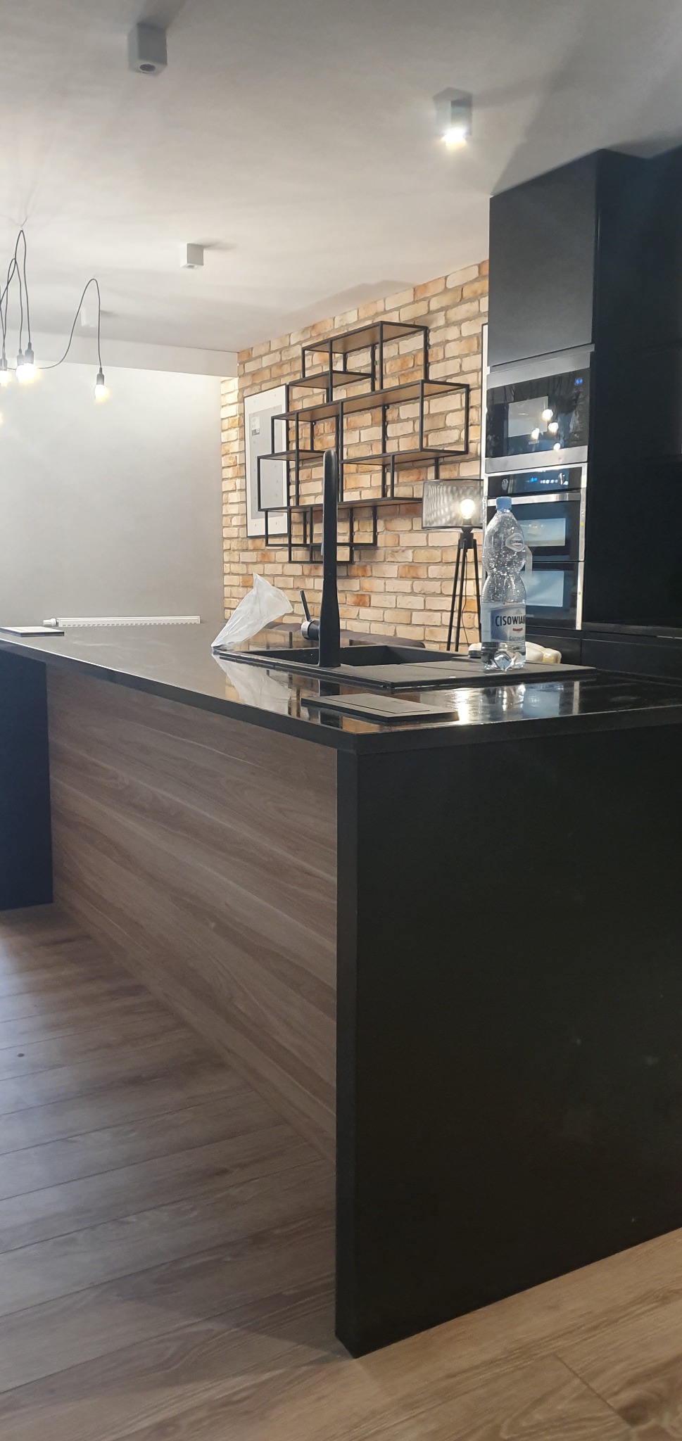 Nowoczesna kuchnia z wyspą, czarnym blatem i frontami z drewnopodobnego laminatu, ceglana ściana z metalowym regałem w tle, widoczna butelka wody mineralnej na blacie.