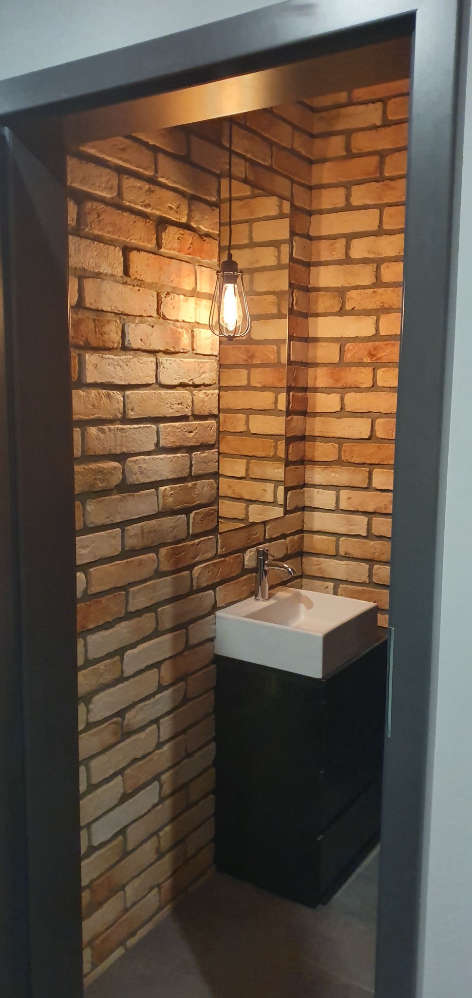 Niewielka łazienka z umywalką nablatową, czarną szafką i ścianami wyłożonymi cegłą, oświetlona wiszącą lampą z żarówką Edisona.