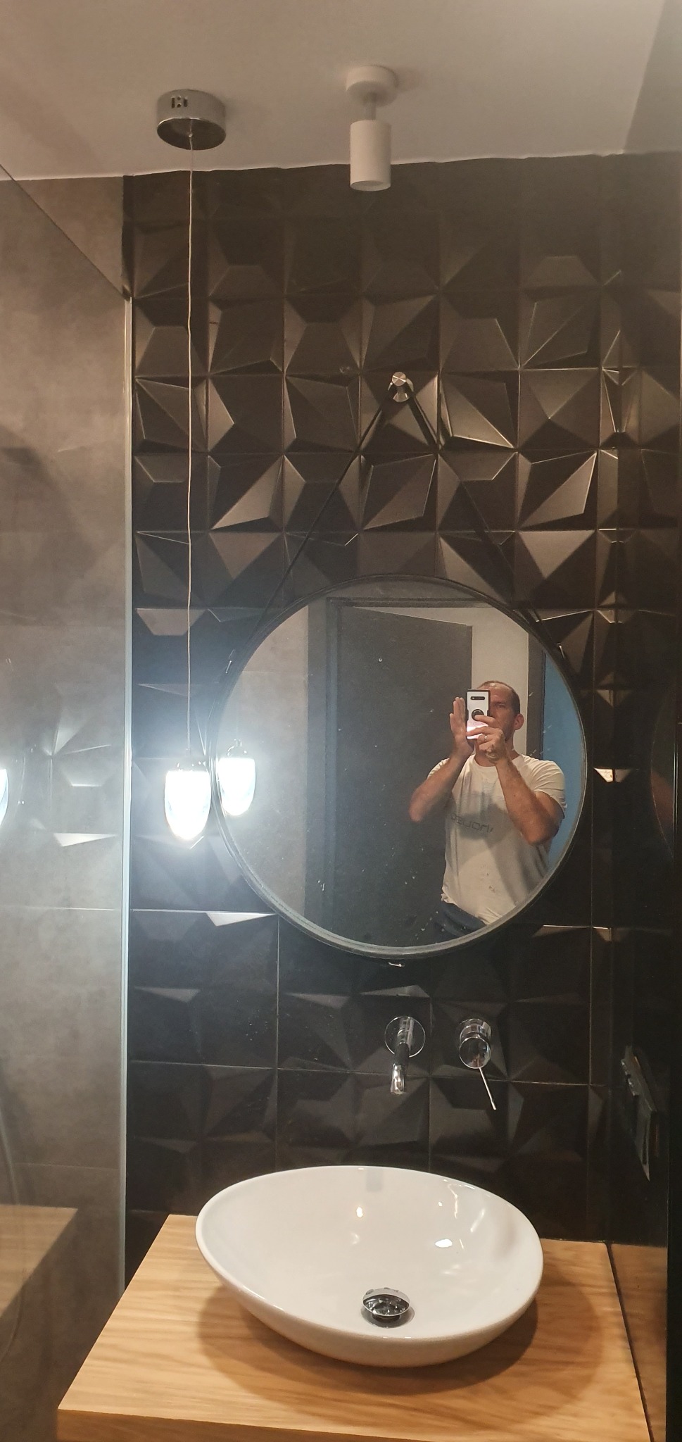 Nowoczesna łazienka z umywalką nablatową, czarnymi trójwymiarowymi płytkami ściennymi i okrągłym lustrem, w którym odbija się osoba robiąca zdjęcie telefonem.