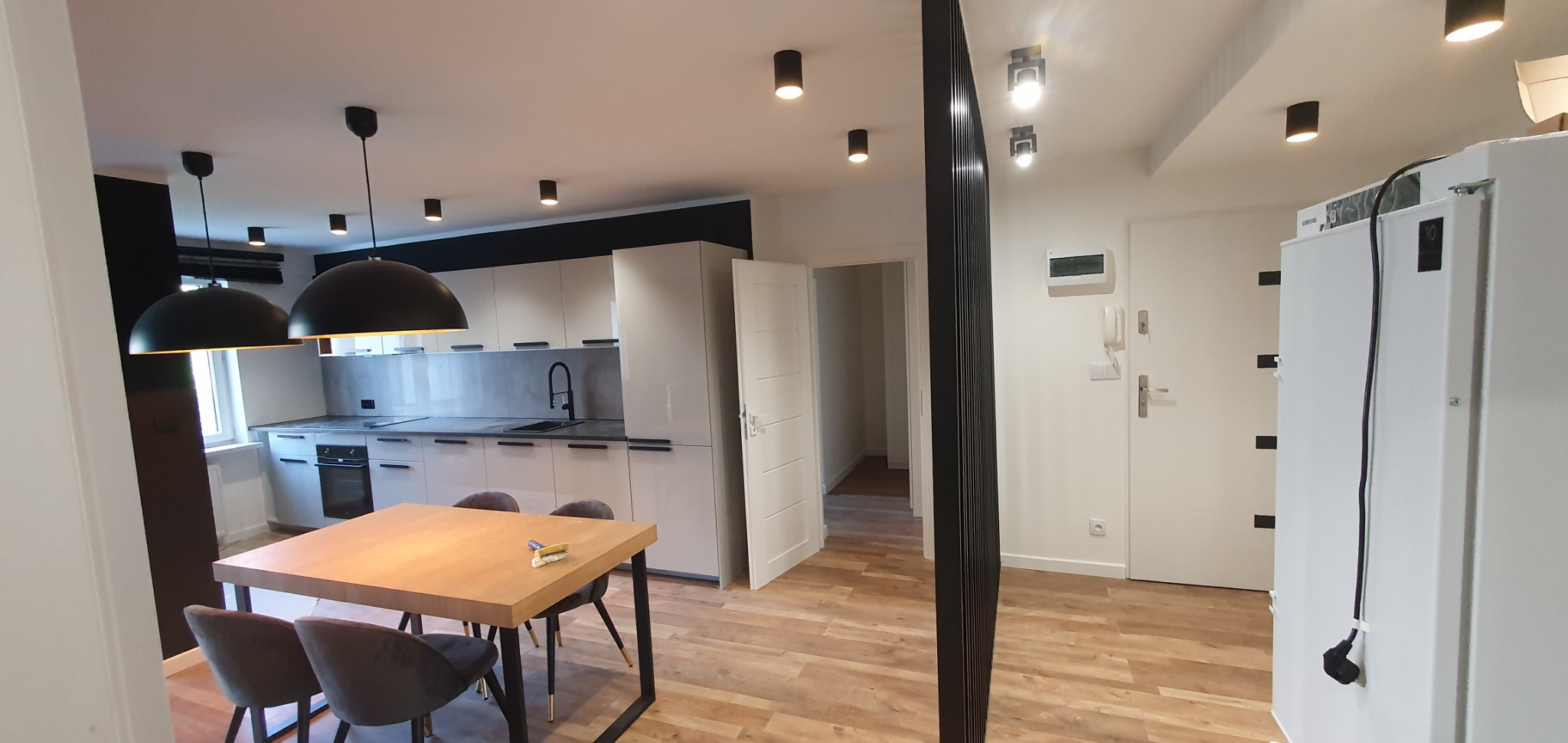 Jasna kuchnia z białymi szafkami, szarym blatem i czarnymi akcentami, widok na stół jadalny z szarymi krzesłami, drewniana podłoga, czarne lampy wiszące i sufitowe.