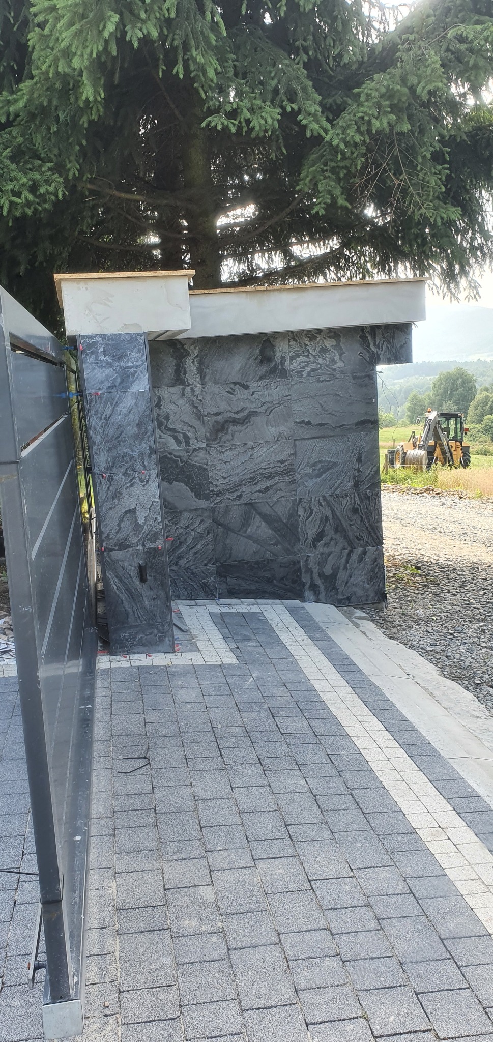 Fragment ogrodzenia z czarnych płyt kamiennych o wyraźnej fakturze, z widocznymi elementami montażowymi, na tle kostki brukowej i krajobrazu z koparką w oddali.