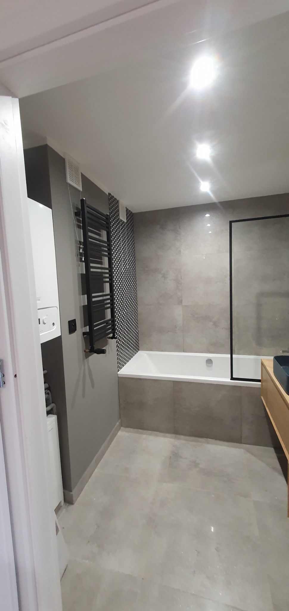 Nowoczesna łazienka w odcieniach szarości z białą wanną, czarnym grzejnikiem drabinkowym i umywalką na drewnianej szafce. W tle widoczny bojler i fragment pralki za otwartymi drzwiami.