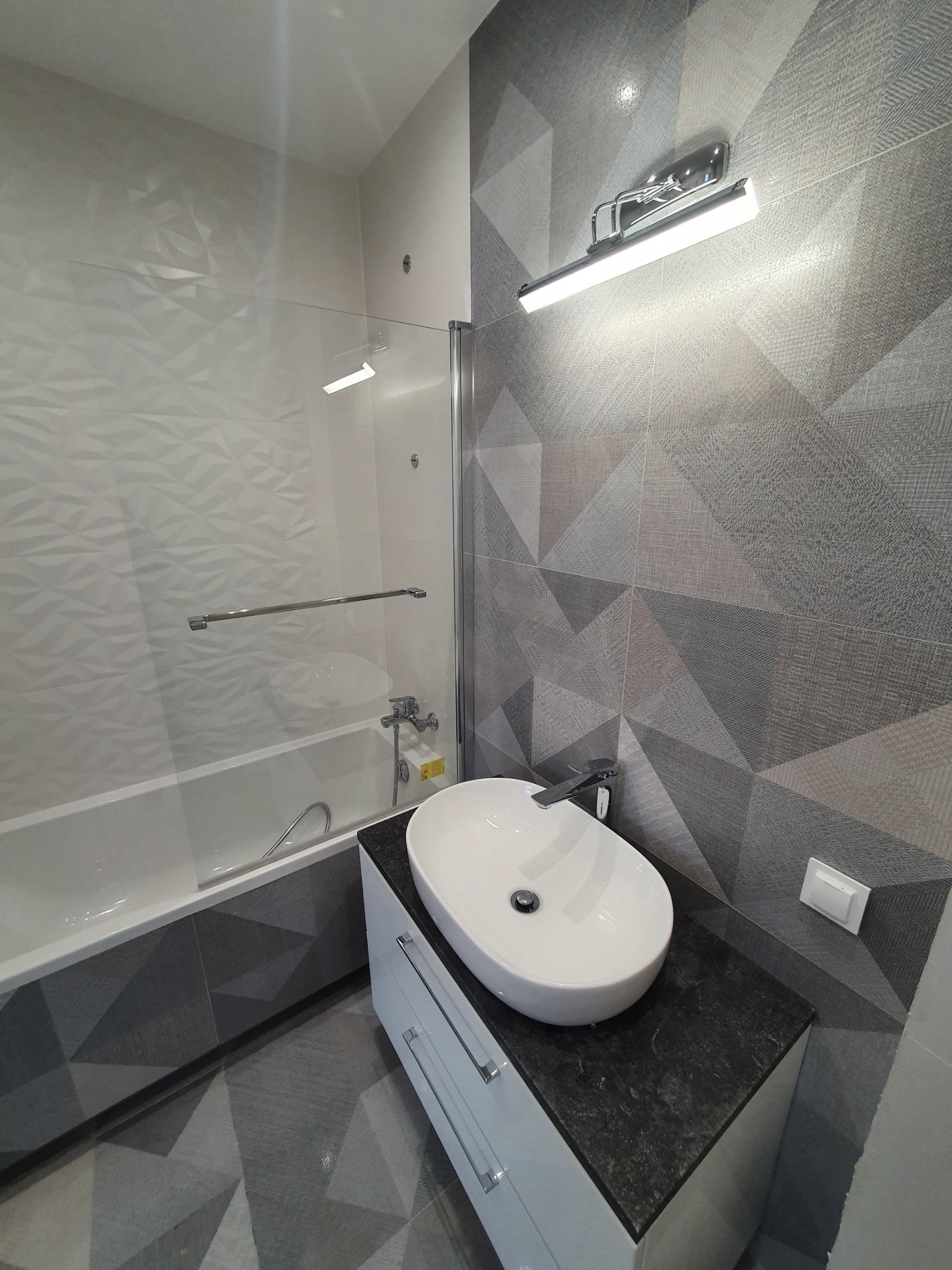 Nowoczesna łazienka z geometrycznymi płytkami w odcieniach szarości, owalna umywalka nablatowa na białej szafce z ciemnym blatem, przeszklona kabina nawannowa, oświetlenie punktowe nad umywalką.