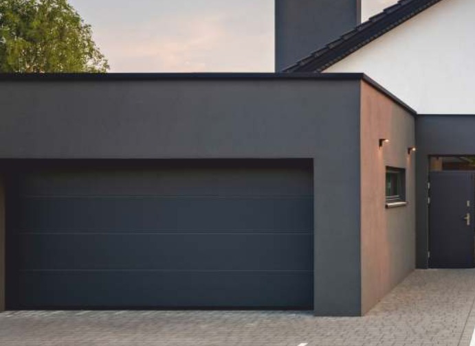 Nowoczesny, antracytowy garaż z segmentową bramą, oświetlony dwoma kinkietami, przylegający do domu z szarą elewacją i małym oknem.