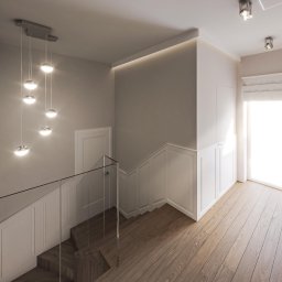 AGNIESZKA PIEC - Jasne wnętrze domu z drewnianą podłogą, schodami z szklaną balustradą i wiszącą lampą kulistą, oświetlenie LED w suficie, obraz na ścianie