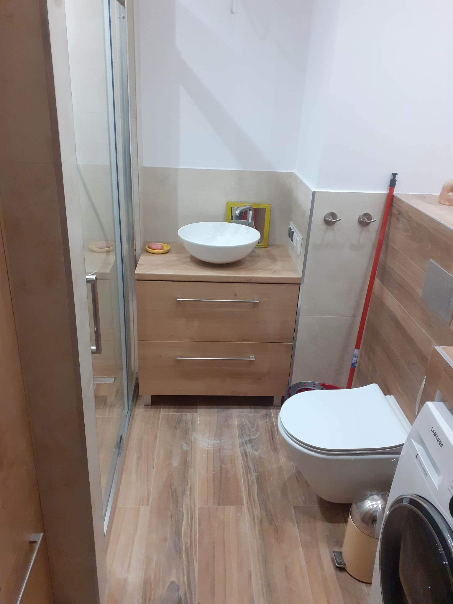 Umywalka nablatowa w okrągłym kształcie na drewnianej szafce z dwiema szufladami, obok kabina prysznicowa ze szklanymi drzwiami, toaleta wisząca oraz pralka w łazience wykończonej płytkami...