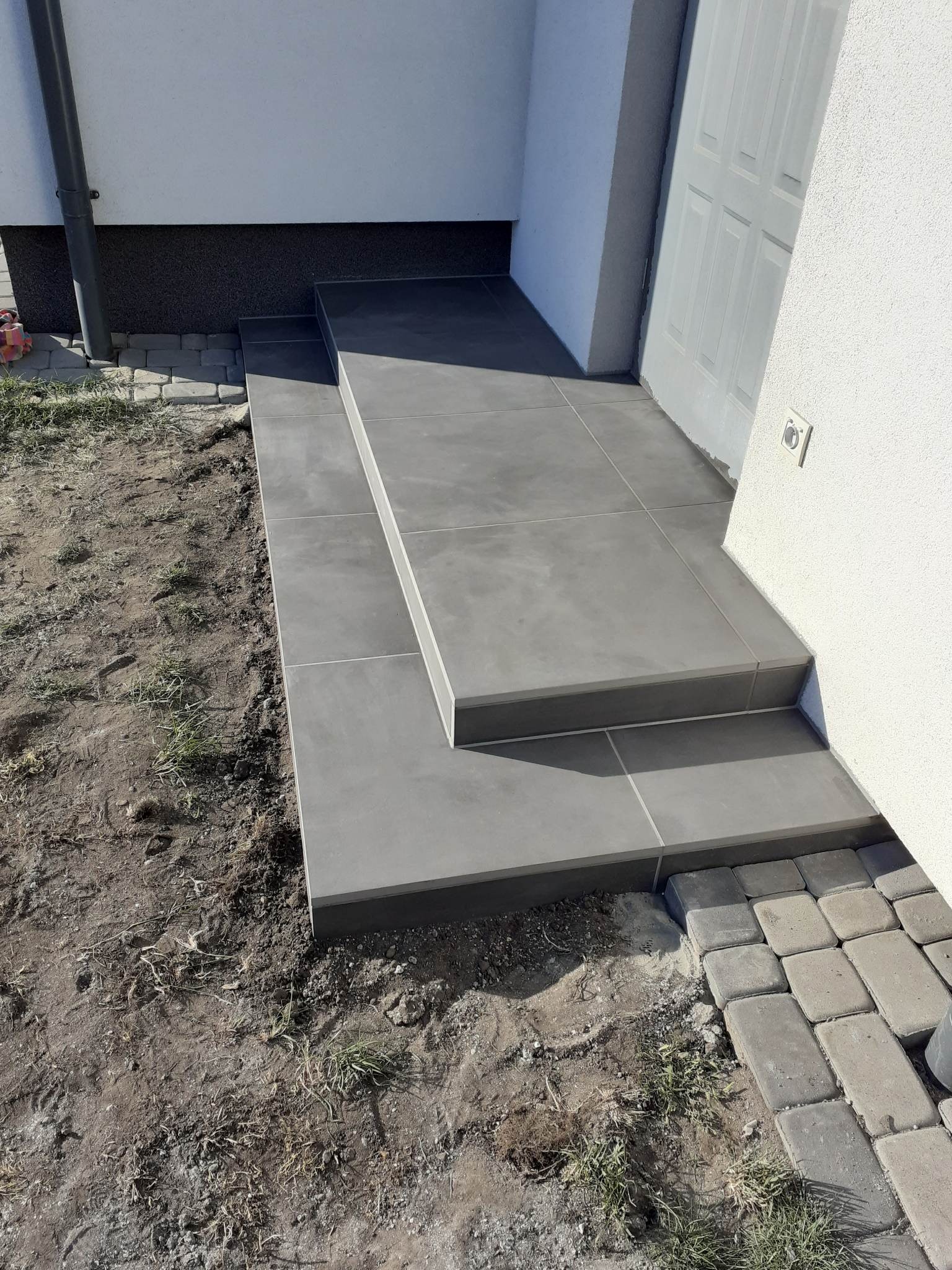 Schody wejściowe do domu wyłożone szarymi płytkami ceramicznymi, z widocznym fragmentem elewacji i kostki brukowej.