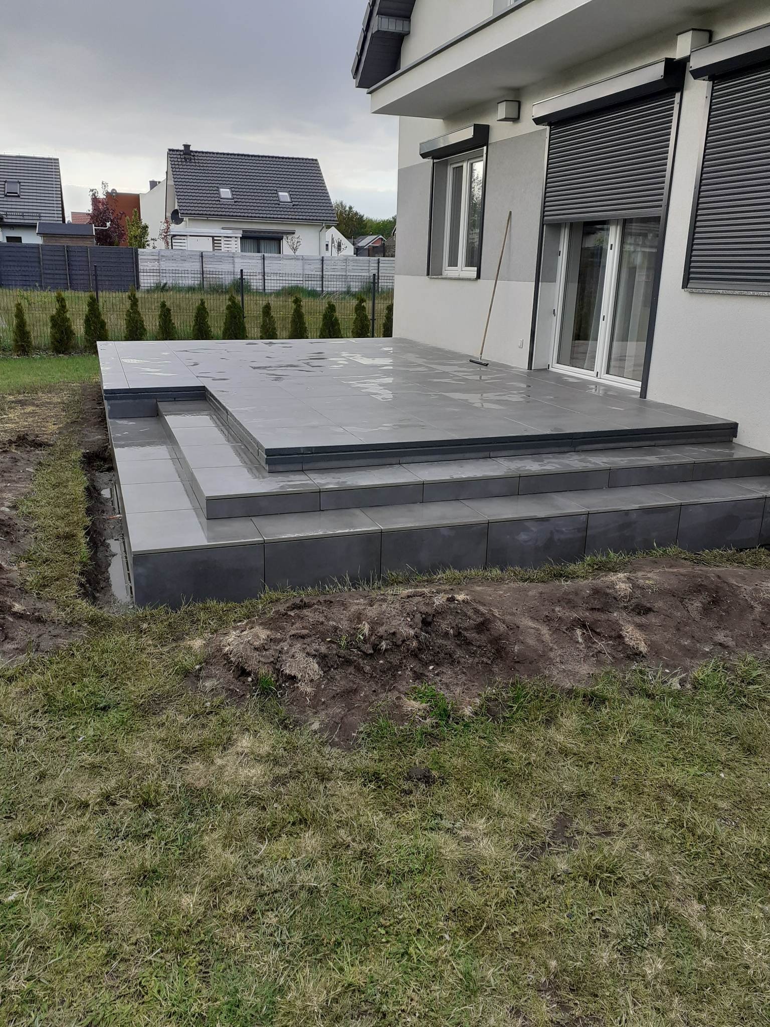 Nowoczesny taras z szarych płytek ceramicznych, wykończony schodami, z widocznym fragmentem trawnika i domu w tle.
