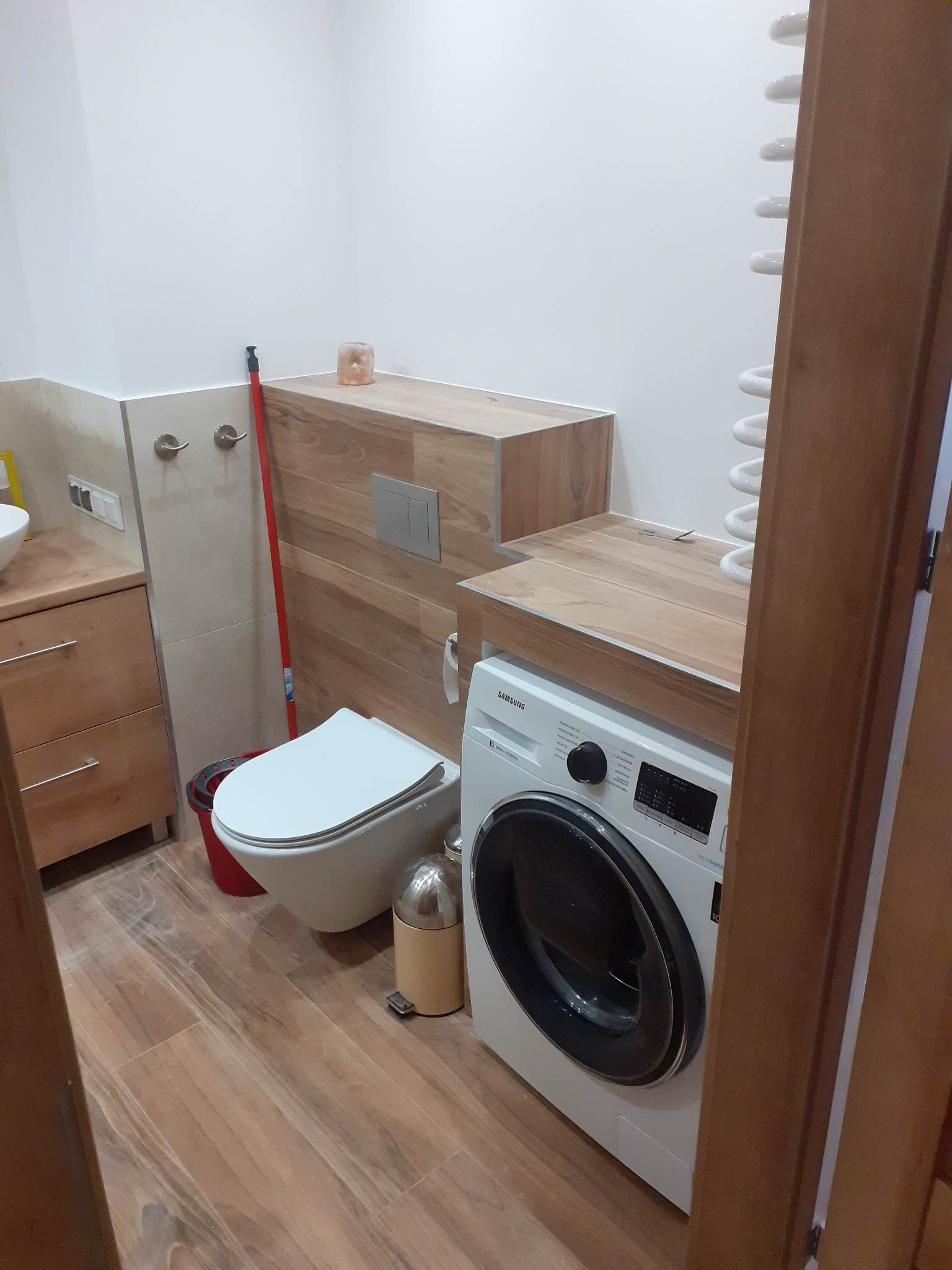 Wykończona łazienka z toaletą, pralką Samsung i drewnopodobnymi płytkami na podłodze i zabudowie, widok zza futryny drzwi.