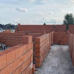Wznoszone ściany z czerwonej cegły ceramicznej na budowie domu jednorodzinnego, widoczne stalowe pręty zbrojeniowe wystające ponad mur, w tle zabudowa osiedlowa pod błękitnym niebem.