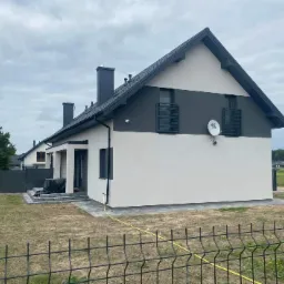 Nowoczesny, dwukondygnacyjny dom jednorodzinny z szarą elewacją w górnej części i białą w dolnej, widoczny zza czarnego metalowego ogrodzenia. Na ścianie zamontowana antena satelitarna 4K. W tle...